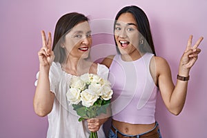 Hispanic mother and daughter holding bouquet of white flowers smiling looking to the camera showing fingers doing victory sign