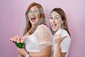 Hispanic mother and daughter holding pink tulips bouquet pointing thumb up to the side smiling happy with open mouth