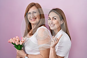 Hispanic mother and daughter holding pink tulips bouquet smiling happy pointing with hand and finger to the side