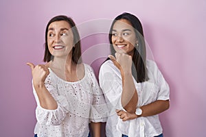 Hispanic mother and daughter together smiling with happy face looking and pointing to the side with thumb up