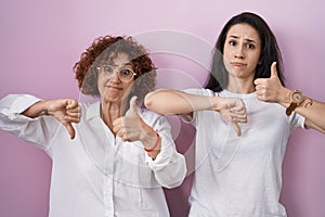 Hispanic mother and daughter wearing casual white t shirt over pink background doing thumbs up and down, disagreement and