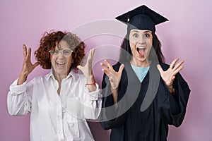 Hispanic mother and daughter wearing graduation cap and ceremony robe celebrating crazy and amazed for success with arms raised