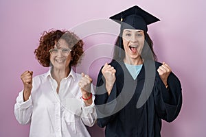 Hispanic mother and daughter wearing graduation cap and ceremony robe celebrating surprised and amazed for success with arms