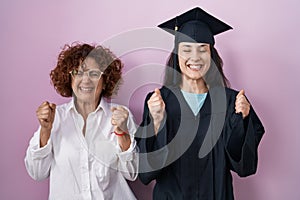 Hispanic mother and daughter wearing graduation cap and ceremony robe excited for success with arms raised and eyes closed