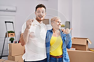 Hispanic mother and son holding keys of new home pointing thumb up to the side smiling happy with open mouth