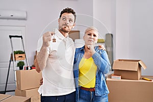 Hispanic mother and son holding keys of new home serious face thinking about question with hand on chin, thoughtful about