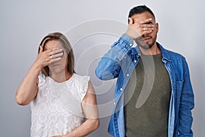 Hispanic mother and son standing together covering eyes with hand, looking serious and sad