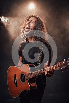 Hispanic musician emotionally singing and playing guitar in stage lights, surrounded by smoke