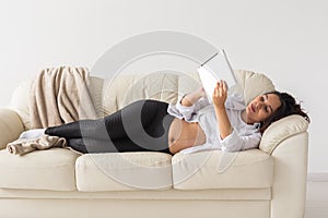 Hispanic pregnant woman using tablet computer while lying on sofa at home. Pregnancy and information for parenthood