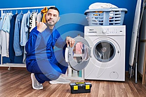 Hispanic repairman working on washing machine pointing thumb up to the side smiling happy with open mouth