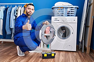 Hispanic repairman working on washing machine smiling happy and positive, thumb up doing excellent and approval sign
