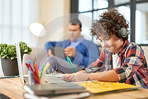 Hispanic school boy wearing headphones, preparing homework while sitting at the desk at home. Father and son spending