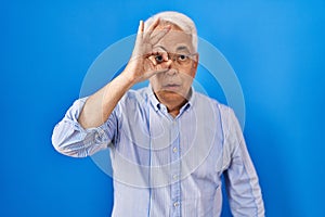 Hispanic senior man wearing glasses doing ok gesture shocked with surprised face, eye looking through fingers