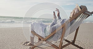 Hispanic senior woman relaxing on sunbed on beach at sunset, using smartphone