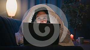 Hispanic smiling happy woman Arabian girl using computer browsing laptop at night at home under blanket game addiction