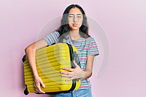 Hispanic teenager girl with dental braces holding cabin bag puffing cheeks with funny face