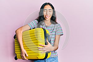 Hispanic teenager girl with dental braces holding cabin bag sticking tongue out happy with funny expression