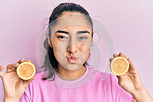 Hispanic teenager girl with dental braces holding lemon puffing cheeks with funny face