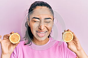 Hispanic teenager girl with dental braces holding lemon sticking tongue out happy with funny expression
