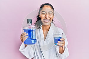 Hispanic teenager girl with dental braces holding mouthwash sticking tongue out happy with funny expression