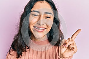 Hispanic teenager girl with dental braces showing orthodontic brackets smiling happy pointing with hand and finger to the side