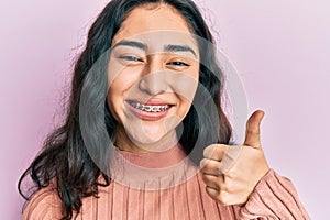 Hispanic teenager girl with dental braces showing orthodontic brackets smiling happy and positive, thumb up doing excellent and