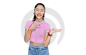 Hispanic teenager girl with dental braces wearing casual clothes amazed and smiling to the camera while presenting with hand and