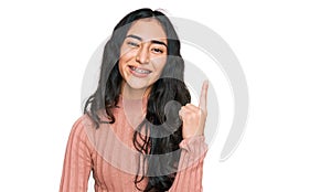 Hispanic teenager girl with dental braces wearing casual clothes with a big smile on face, pointing with hand and finger to the