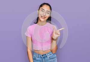 Hispanic teenager girl with dental braces wearing casual clothes with a big smile on face, pointing with hand finger to the side