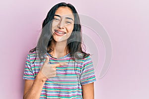 Hispanic teenager girl with dental braces wearing casual clothes cheerful with a smile of face pointing with hand and finger up to