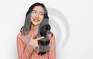 Hispanic teenager girl with dental braces wearing casual clothes cheerful with a smile of face pointing with hand and finger up to