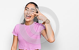 Hispanic teenager girl with dental braces wearing casual clothes doing peace symbol with fingers over face, smiling cheerful