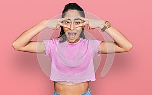 Hispanic teenager girl with dental braces wearing casual clothes doing peace symbol with fingers over face, smiling cheerful