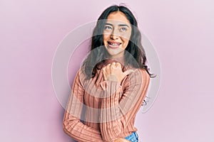 Hispanic teenager girl with dental braces wearing casual clothes smiling with happy face looking and pointing to the side with