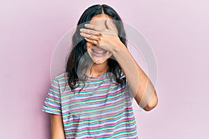 Hispanic teenager girl with dental braces wearing casual clothes smiling and laughing with hand on face covering eyes for surprise