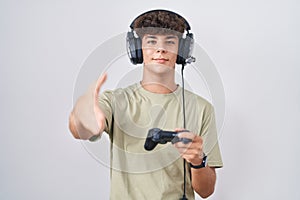Hispanic teenager playing video game holding controller smiling friendly offering handshake as greeting and welcoming