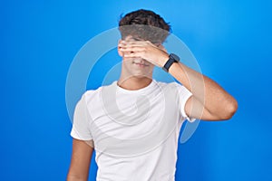 Hispanic teenager standing over blue background covering eyes with hand, looking serious and sad