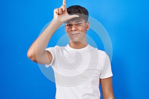 Hispanic teenager standing over blue background making fun of people with fingers on forehead doing loser gesture mocking and