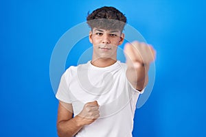 Hispanic teenager standing over blue background punching fist to fight, aggressive and angry attack, threat and violence