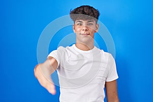 Hispanic teenager standing over blue background smiling friendly offering handshake as greeting and welcoming