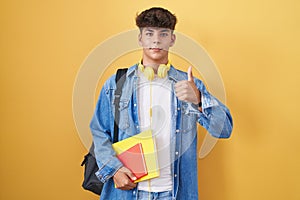 Hispanic teenager wearing student backpack and holding books doing happy thumbs up gesture with hand