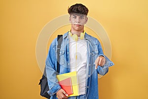 Hispanic teenager wearing student backpack and holding books pointing down looking sad and upset, indicating direction with
