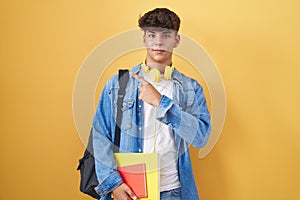 Hispanic teenager wearing student backpack and holding books pointing with hand finger to the side showing advertisement, serious