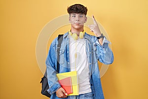 Hispanic teenager wearing student backpack and holding books smiling amazed and surprised and pointing up with fingers and raised