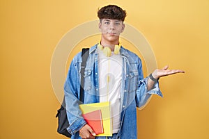 Hispanic teenager wearing student backpack and holding books smiling cheerful presenting and pointing with palm of hand looking at