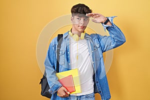 Hispanic teenager wearing student backpack and holding books very happy and smiling looking far away with hand over head