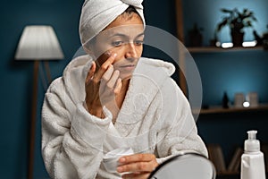 Hispanic woman apply facial cream look in small mirror, healthy lady with towel on head put moisturizing on moisturized