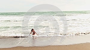 Hispanic woman in bikini standing on sandy beach with cold wind and waves
