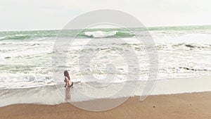 Hispanic woman in bikini standing on sandy beach with cold wind and waves