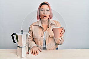 Hispanic woman with pink hair drinking a cup of italian coffee scared and amazed with open mouth for surprise, disbelief face